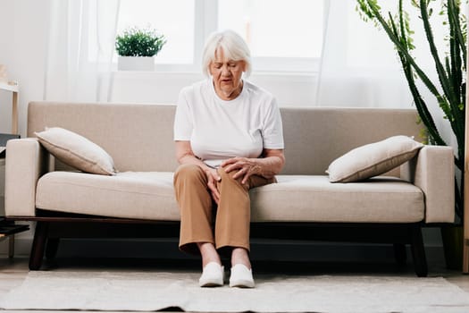 Elderly woman severe pain in her leg sitting on the couch, health problems in old age, poor quality of life. Grandmother with gray hair holds on to her sore knee, problems with joints and ligaments. . High quality photo