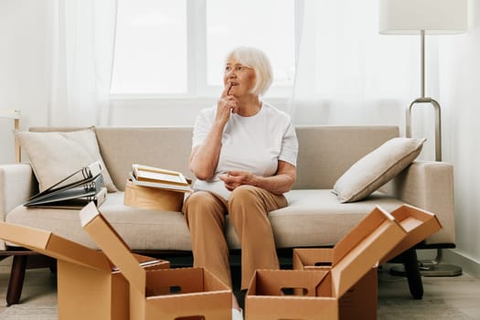 an elderly woman sits on a sofa at home with boxes. collecting things with memories and moving. High quality photo