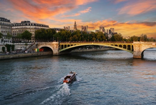 View of the Seine River in Paris at sunset with a bridge in the background, orange clouds on blue sky and a small boat sailing on the river.