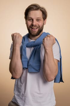 Happy joyful caucasian man shouting, raising fists in gesture I did it, celebrating success, winning, birthday, lottery jackpot goal achievemen, good news. Young adult guy on beige studio background