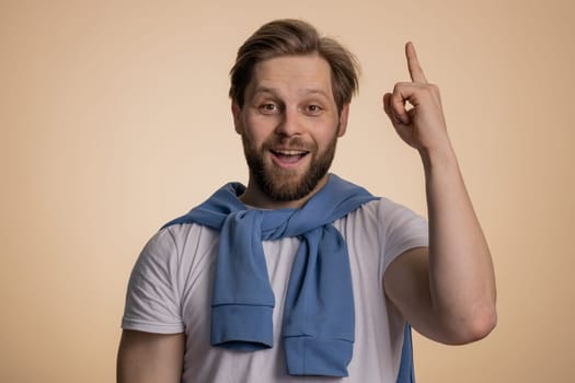 Eureka. Inspired young caucasian man pointing finger up with open mouth, having good idea, plan, startup, showing inspiration motivation gesture, problem solution. Bearded guy on beige background