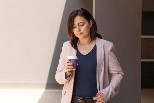 Puzzled Mature 40 yo Woman, Manager Looks At Coffee Cup, Thinking Worryingly Near Office Outdoor, Factory on Background. Caucasian Burnette Female Wears Pink Jacket, Blue Shirt.Horizontal, Copy Space.
