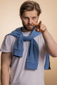 I am watching you. Young confident caucasian man pointing at his eyes and camera, show I am watching you gesture, spying on someone. Handsome bearded guy isolated alone on beige studio background