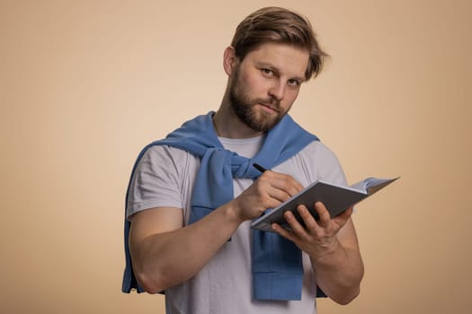 Thoughtful journalist caucasian man making notes, writing down thoughts with pen into notepad notebook diary, to do list, good idea. Handsome bearded guy isolated alone on beige studio background