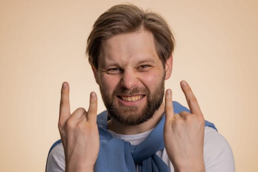 Overjoyed caucasian man showing rock n roll gesture by hands, cool sign, shouting yeah with crazy expression, dancing, emotionally rejoicing in success. Young happy guy isolated on beige background