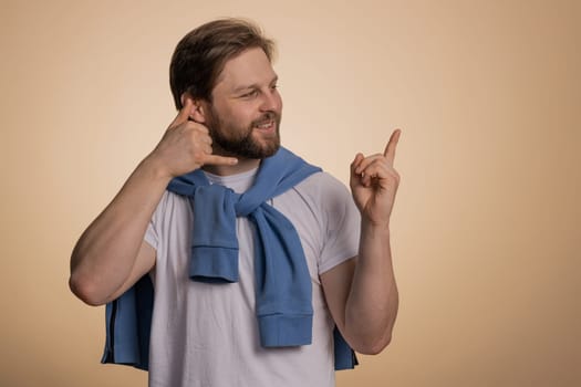 Call us, here is contact number. Handsome bearded caucasian man in t-shirt looking at camera doing phone gesture like says hey you call me back conversation. Guy isolated on beige background, indoor