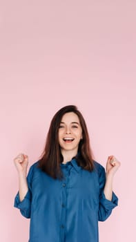 Euphoric Woman Celebrating Success, Raising Fists in Triumph: Isolated Pink Background Portrait. Joyful Woman Celebrating Achievement, Raising Fists in Excitement: Isolated Pink Background Portrait.