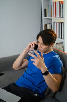 Young man freelancer sitting on couch at home and talking on mobile phone. People, technology and communication.