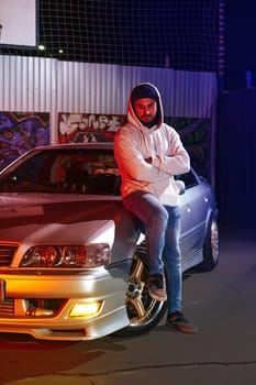 A young man standing next to his sport car at night parking in neon light