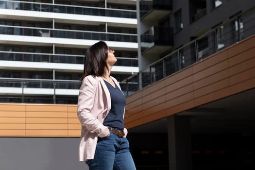 Happy Mature 40 yo Woman Enjoys New Apartment Campus, Complex In Sunny Day. Female Brunette Wears Casual Cloth, Looks At Construction, Modern Residential Building, Standing on Parking. Horizontal.