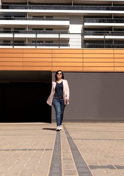Happy Mature 40 yo Woman Walks In Parking Of New Apartment Campus, Complex In Sunny Day. Female Brunette Wears Casual Cloth. Construction, Modern Residential Building. Vertical Plane.