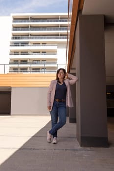 Caucasian Woman Leans To Wall In Parking Area Of New Apartment Campus, Complex In Sunny Day. Female Brunette Wears Casual Cloth. Construction, Modern Residential Building. Vertical Plane. High quality photo