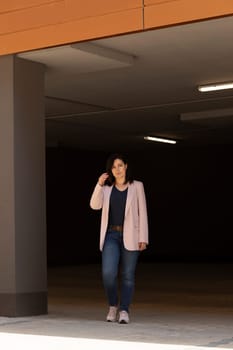 Beautiful Caucasian Woman Walks In Parking Of New Apartment Campus, Complex In Sunny Day. Female Brunette Wears Casual Cloth. Construction, Modern Residential Building. Vertical Plane.