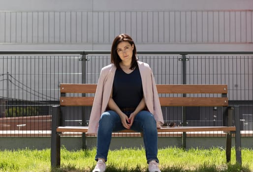 Beautiful Brunette Mature Woman Sitting On A Bench In Residential Campus. Confident Female Is Dressed In Casual Clothes. Rest And Relaxation. Horizontal Plane. High quality photo