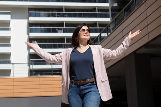 Happy Mature Woman With Open Gesture, Arms Enjoys Modern Residential Complex, Standing In New Apartment Campus In Sunny Day. Female Brunette Wears Casual Cloth, Horizontal, Real Estate Development.