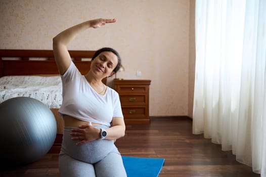 Attractive happy pregnant woman touching her big belly on late pregnancy, stretching her arm, sitting in hero pose on a yoga mat, practicing prenatal fitness for wellness and healthy. Maternity leave