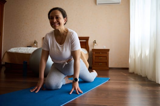 Smiling middle aged multi ethnic pregnant woman in white t-shirt ad gray leggings, exercising on blue yoga mat at home. Prenatal stretching. Sport. Fitness. Healthy active pregnancy lifestyle concept