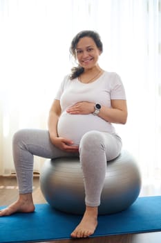 Lovely pregnant woman sitting on fitball, holding hands on her belly, smiling looking at camera, expressing positive emotions, enjoying prenatal fitness and healthy active lifestyle in pregnancy time