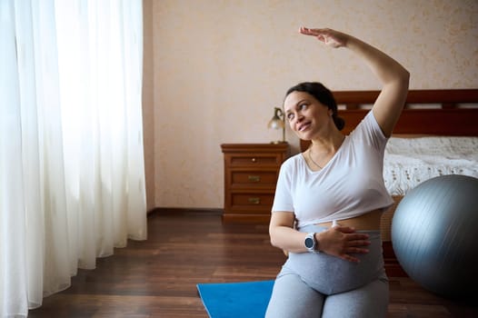 Delightful gorgeous pregnant woman holds her hand on her big bell, stretching her arm right, sitting in hero pose on a yoga mat, practicing prenatal fitness for wellness and healthy in pregnancy time