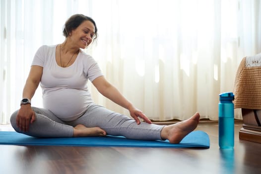 Smiling happy pregnant woman doing prenatal stretching exercises on yoga mat at home. Gravid expectant mother reaching arm to her leg, stretching body for wellness in pregnancy time