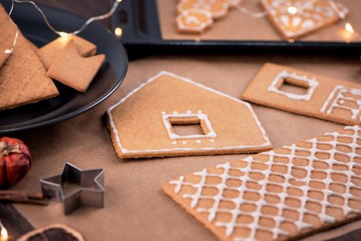 Woman is decorating gingerbread cookies house with white frosting icing cream topping on wooden table background, baking paper in kitchen, close up, macro.