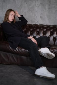 beautiful confident brunette woman in black on a brown leather couch in grey dark room. business woman. feminine. millennial people.