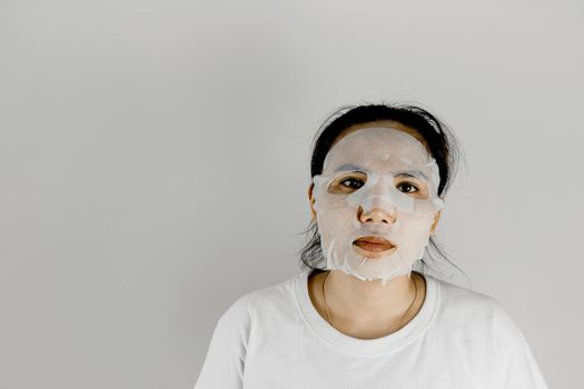 Front view of Asian woman wearing white t-shirt and covering her face with a sheet mask.