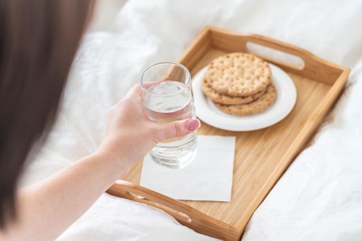 Female hand holding glass of water over tray with crackers on a bed