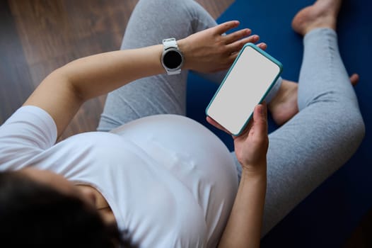 Top view pregnant woman checks smart watch to control pulse and heart rate, relaxing with smartphone on yoga mat after training at home, browsing mobile application, resting after meditation practice