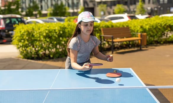 Young teenager girl playing ping pong. She holds a ball and a racket in her hands. Playing table tennis outdoors in the yard.
