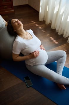 Overhead view beautiful pregnant woman, relaxing with her eyes closed, leaning on a fitball, holding hands on her belly, sitting on a mat at home. Daylight. Prenatal fitnesss and relaxation exercises