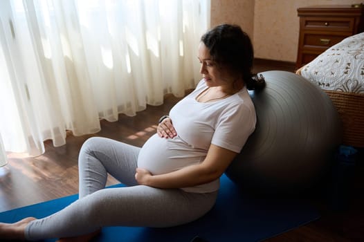 Beautiful pregnant woman, expectant gravid mother caressing her belly, relaxing on a yoga mat leaning on fit ball after, enjoying her pregnancy and maternity lifestyle in a tranquil homely atmosphere