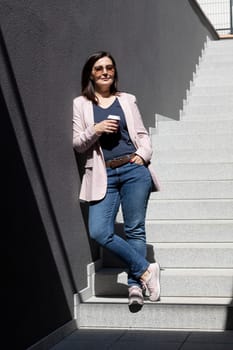 Smiling Beautiful Brunette Woman Hold Paper Cup Of Coffee Standing On Stairs Near Office Outdoor, Dressed In Stylish Casual Clothes, Summer Sunny Day. Taking A Break. Lifestyle Portrait Mature Female