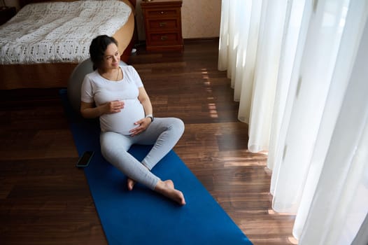 Top view of delightful relaxed pregnant woman in white t-shirt, sitting barefoot on a blue yoga mat, stroking her big belly, relaxing after prenatal relaxation exercises at home. Healthy pregnancy