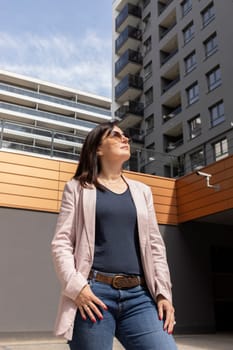 Pretty Mature 40 yo Woman Enjoys View Of New Apartment Campus, Complex In Daytime. Female Brunette Wears Casual Cloth, Looks At Construction, Modern Residential Building, Vertical Plane.