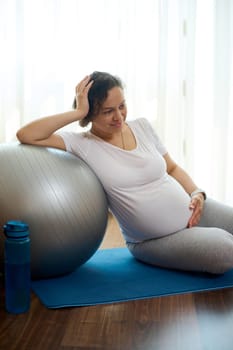 Gravid woman holding her hand on her pregnant belly, relaxing on exercise mat after prenatal yoga practice and meditation, expressing positive emotions of a healthy active lifestyle in pregnancy time