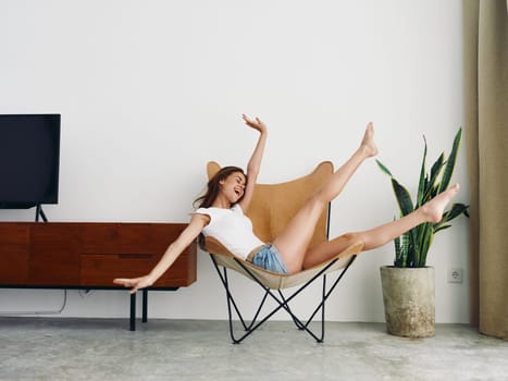 Woman lying in a leather armchair raised arms and legs cheerful happiness and a smile, relaxing at home stylish modern interior with white walls, copy space. High quality photo