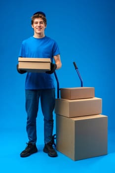 Young happy delivery man carrying boxes on truck against blue background in studio