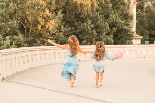 Daughter mother run holding hands. In blue dresses with flowing long hair against the backdrop of a sunset and a white building