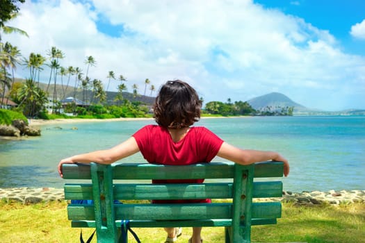 Young woman resting on park bench along Hawaiian ocean, Kokohead Crater in background