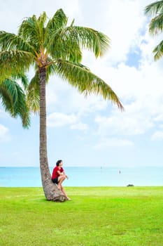 Young woman in casual clothes resting or leaning against one coconut or palm tree on grassy lawn next to Hawaiian ocean
