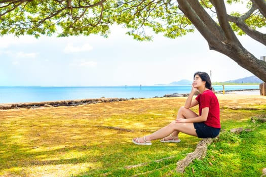 Young woman resting in shade of large canopy tree in Hawaii, with ocean horizon in background