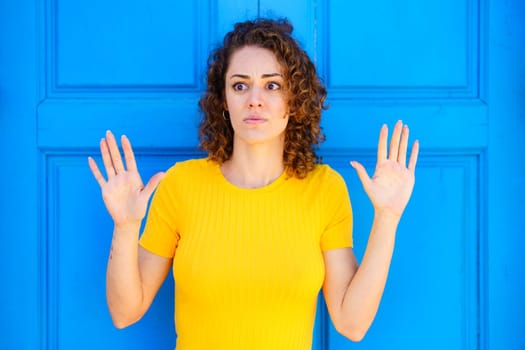 Afraid young female in yellow t shirt with curly brown hair and raised hands looking away against blue wall on street