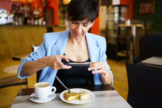 Smiling young female in blue jacket sitting at table, with cup of coffee and looking at screen of mobile phone while taking picture of snack in restaurant