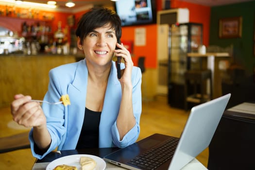 Positive female freelancer in casual clothes smiling while eating and talking on mobile phone at table with laptop in modern cafe indoors