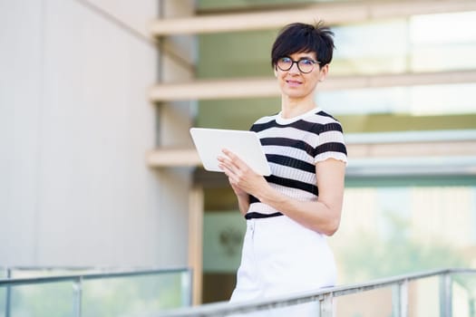 Adult female in glasses and casual clothes looking at camera while standing on balcony of modern building with tablet