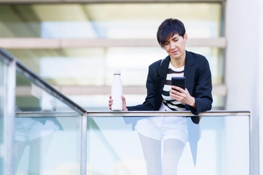 Serious entrepreneur in smart casual clothes standing near glass wall and browsing mobile phone while holding bottle of fresh water