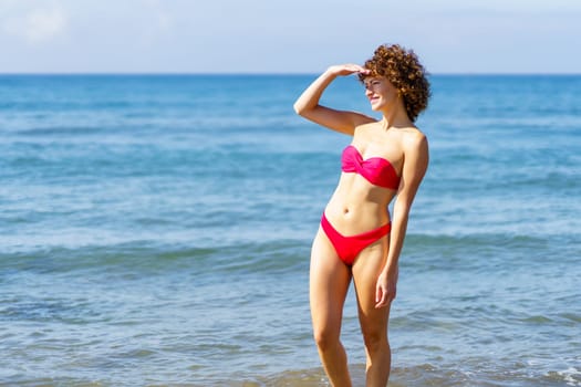 Side view of smiling young female with curly red hair, in bikini standing in seawater with hand at face covering sunlight while looking away against blue wavy sea