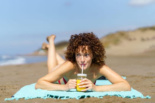 Young female with curly red hair lying on beach and sipping orange juice from glass cup with straw looking away