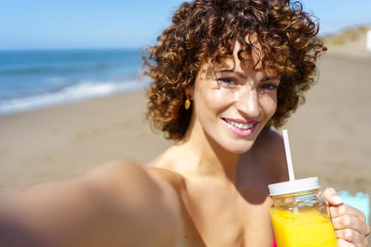Portrait of smiling young woman with curly hair drinking refreshing orange juice while taking selfie on beach during summer vacation